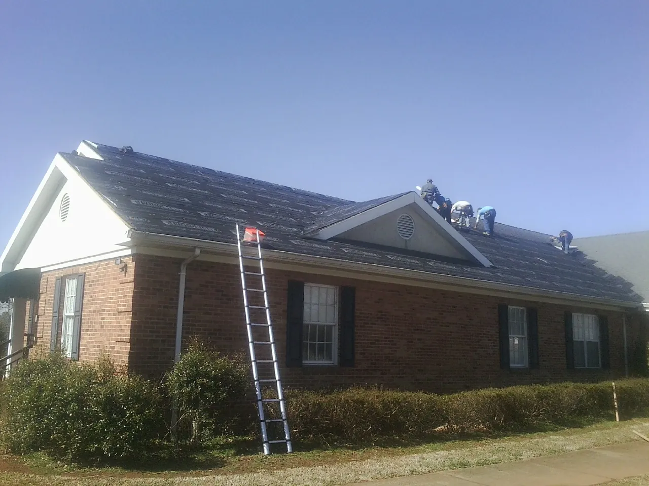 Crew tearing shingles off a brick ranch roof with a ladder set — Metal Roof Repair work by our crew in Wonder Lake