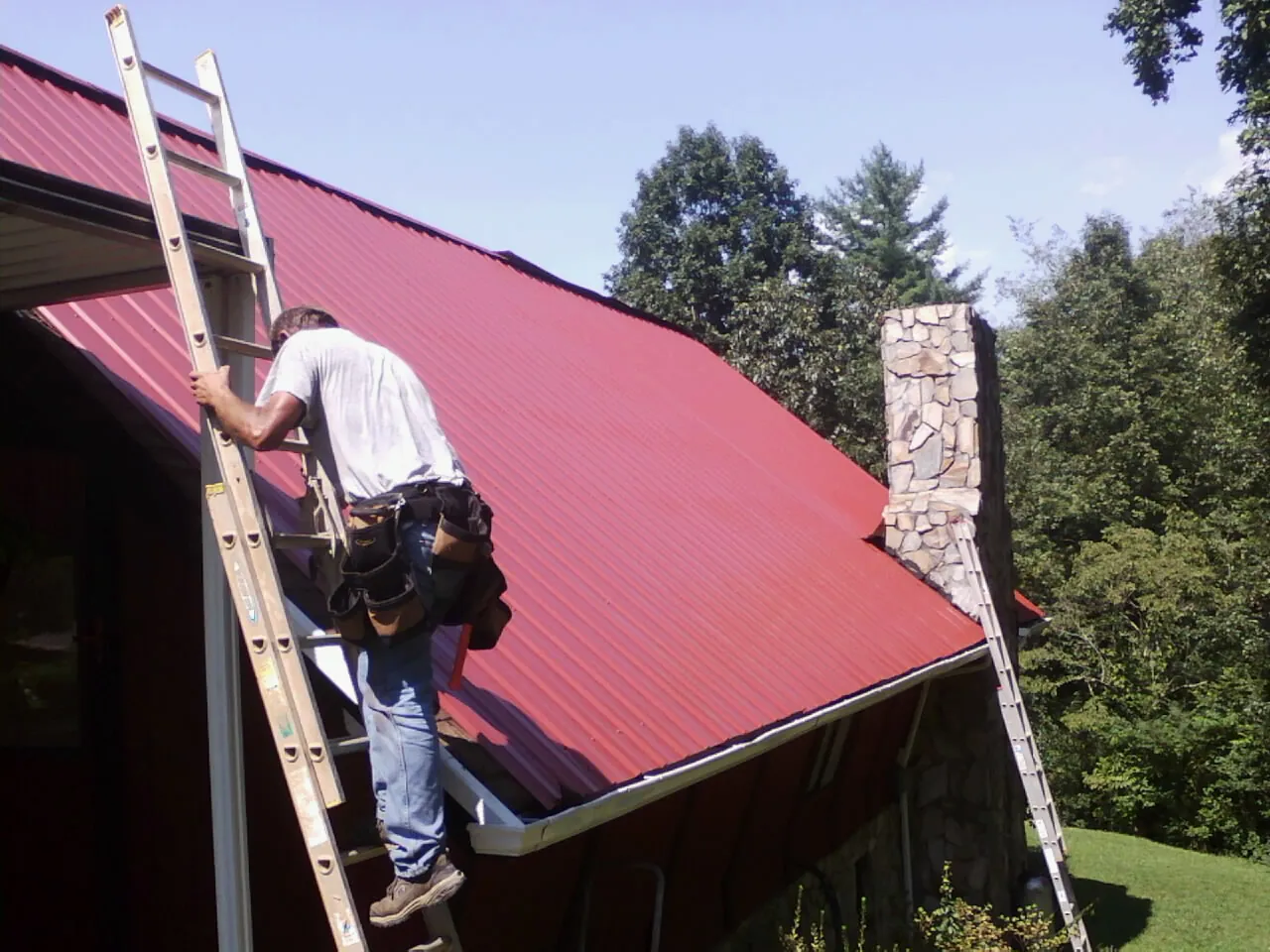 Roofer on a ladder beside a red metal roof and stone chimney — Soffit & Fascia Repair by Roofing Brothers in Wonder Lake